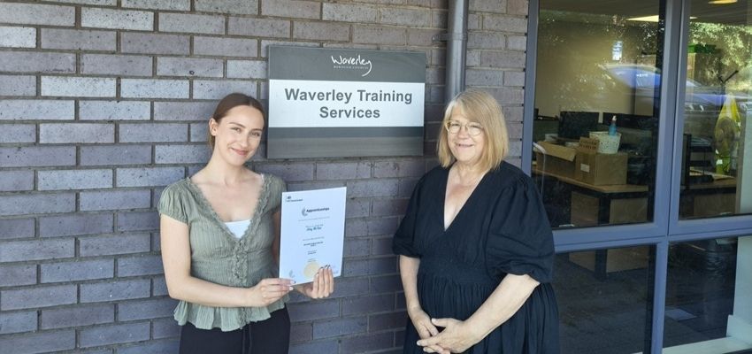 Apprentice Amy holding up a certificate of BAL3 next to colleague outside the Waverley Training Services office.